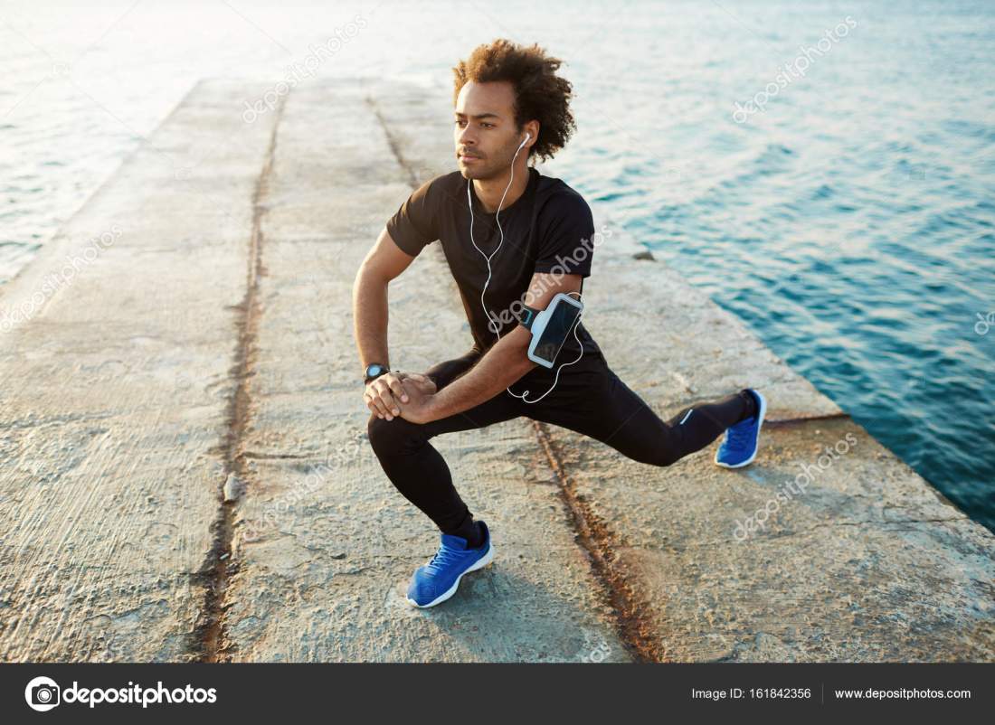 Young male dark-skinned runner with beautiful fit body warming-up his muscles before strength training cardio workout. Man athlete in sportswear stretching legs with lunge hamstring stretch exercise on pier.